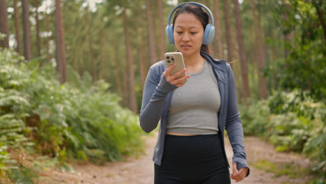 Woman-Wearing-Wireless-Headphones-Exercising-Doing-Work-Out-Outdoors-Streaming-Music-Or-Podcast-From-Mobile-Phone-Running-Along-Track-In-Forest-Wearing-Sports-Clothing-Shot-In-Real-Time